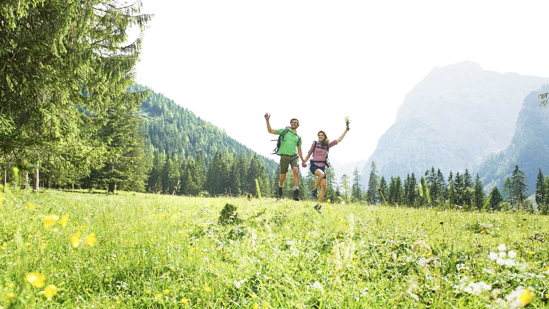 Wanderpaar springt fröhlich über Sommerwiese in Pertisau am Achensee vor alpiner Bergkulisse.